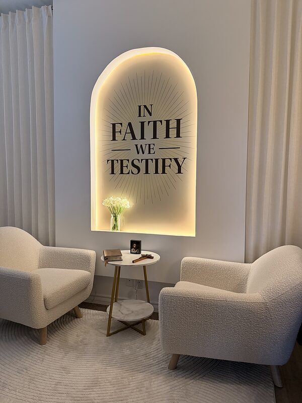 The completed &lsquo;In Faith We Testify&rsquo; set &mdash; two cream armchairs and a small marble table with a Bible and wooden cross beneath the warmly backlit arch carrying the In Faith We Testify wordmark