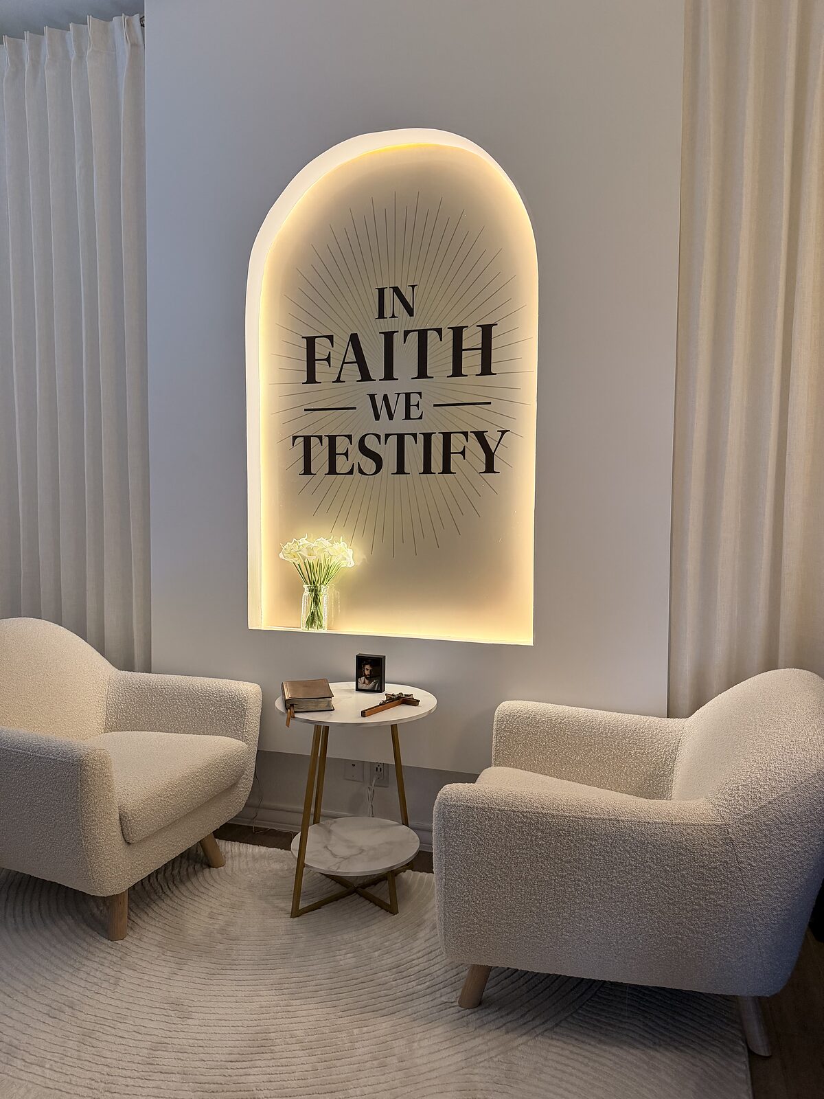 The completed &lsquo;In Faith We Testify&rsquo; set &mdash; two cream bouclé armchairs facing a small marble table holding a Bible and a wooden cross, beneath the warmly backlit arch carrying the In Faith We Testify wordmark, with white calla lilies on the ledge and floor-length ivory curtains on either side
